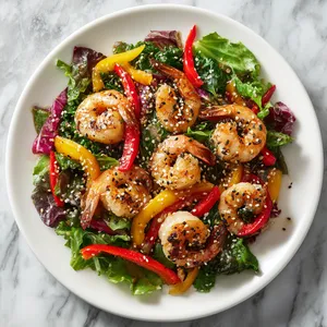 Juicy pink shrimp scattered across a vibrant bed of greens, bell peppers, and a hint of red from the chilies. The whole dish pops against a crisp white plate, topped with a smattering of sesame seeds.