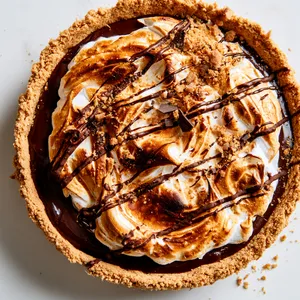 From above, a round tart with a golden graham crust is filled with glossy dark chocolate and topped with swirls of toasted marshmallow meringue, deeply bronzed at the tips, finished with zigzags of chocolate drizzle, graham crumble, and a few tiny shards of chocolate for a striking bakery-style look.