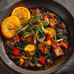 The dish looks vibrant on a deep, warm serving dish. The dark, almost ebony stew, generously speckled with colorful cubes of bell pepper, chunks of roasted plantains, and dark leafy greens. A garnish of bright orange slices and fresh herbs rest casually on the side, providing a burst of natural, lively color.
