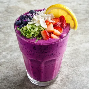 Purple smoothie with an ombre, transitioning from almost pink at the bottom to a deep, beautiful violet hue at the top. Additional toppings of finely chopped green cabbage, strawberries, blueberries, and coconut flakes brings in a cavalcade of colors, textures, and tastes. A vibrant yellow lemon slice rests against the rim of the glass, providing a visually appealing contrast.