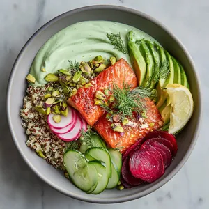 From above, a wide shallow bowl shows a swoosh of pale green yogurt, a neat mound of fluffy quinoa, glossy roasted salmon with crisp edges, ruby beet wedges, cucumber ribbons, avocado fan, radish slices, and a scatter of pistachios, dill, mint, and microgreens for a bright rainbow effect.