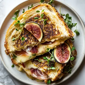 From the top, you see slices of sourdough lightly toasted to golden perfection and drizzled with a glossy fig glaze. A cross-section reveals layers of melted brie, fig jam, and curled prosciutto peeking out from the bread. Served on a clean white plate, the sandwich is garnished with fresh fig slices and sparkling microgreens for a dash of color and freshness.