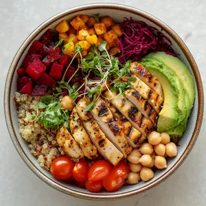 A ceramic bowl filled to the brim with radiantly colorful layers of quinoa, cherry tomatoes, diced avocado, shredded beetroot, boiled chickpeas, and grilled chicken. The bowl is garnished with a flourish of microgreens and finished with a drizzle of tangy vinaigrette, adding a beautiful shine.