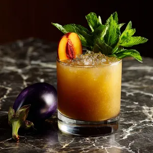 The finished drink exhibits a rich, golden hue due to the peach puree and bourbon, finished with a hint of violet from the eggplant. A mint sprig and a peach slice perched daintily on the rim of the glass add a pop of color, creating a visually stimulating yet sophisticated cocktail.